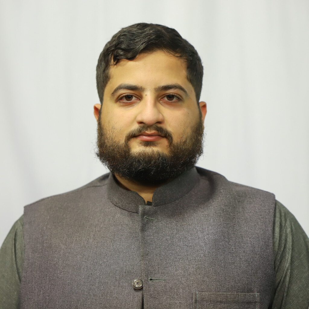 Portrait of a man with dark hair and beard, wearing a gray Nehru-style vest against a light backdrop, looking at the camera.