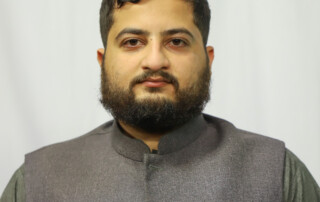 Portrait of a man with dark hair and beard, wearing a gray Nehru-style vest against a light backdrop, looking at the camera.