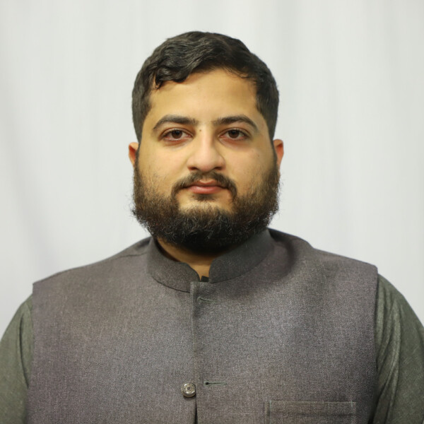 Portrait of a man with dark hair and beard, wearing a gray Nehru-style vest against a light backdrop, looking at the camera.